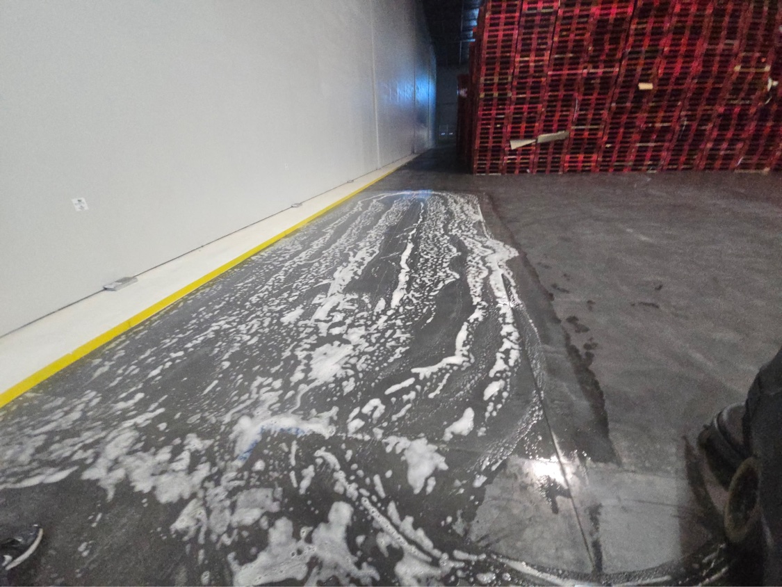 Crew scrubbing and treating a warehouse floor aisle between stacks of red pallets