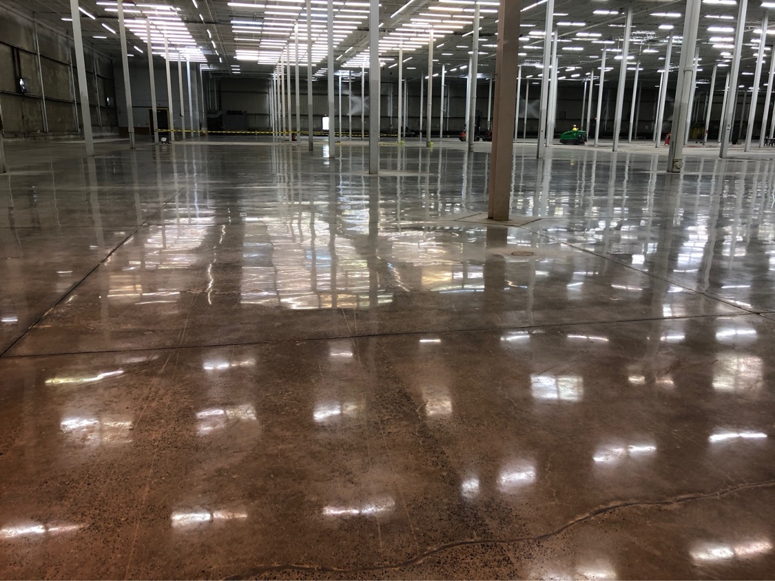 Enormous warehouse floor with a warm brown stained, mirror-polished finish reflecting rows of ceiling lights between the columns